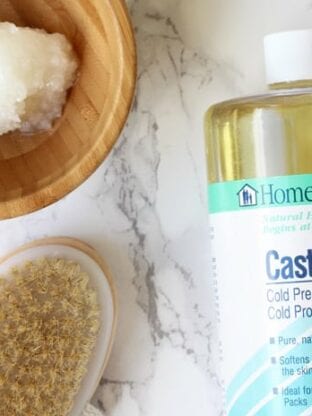 A bottle of castor oil, dry brush, and wooden bowl with oil in it all sitting together on a counter.