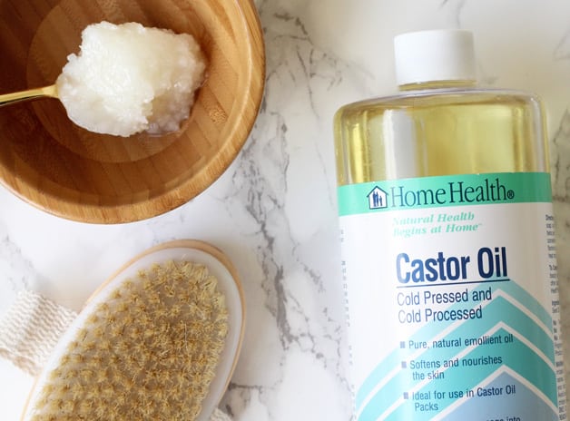 A bottle of castor oil, dry brush, and wooden bowl with oil in it all sitting together on a counter.
