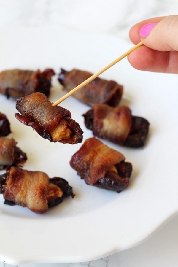 A plate with bacon wrapped dates and with a tooth pick picking one of them up.