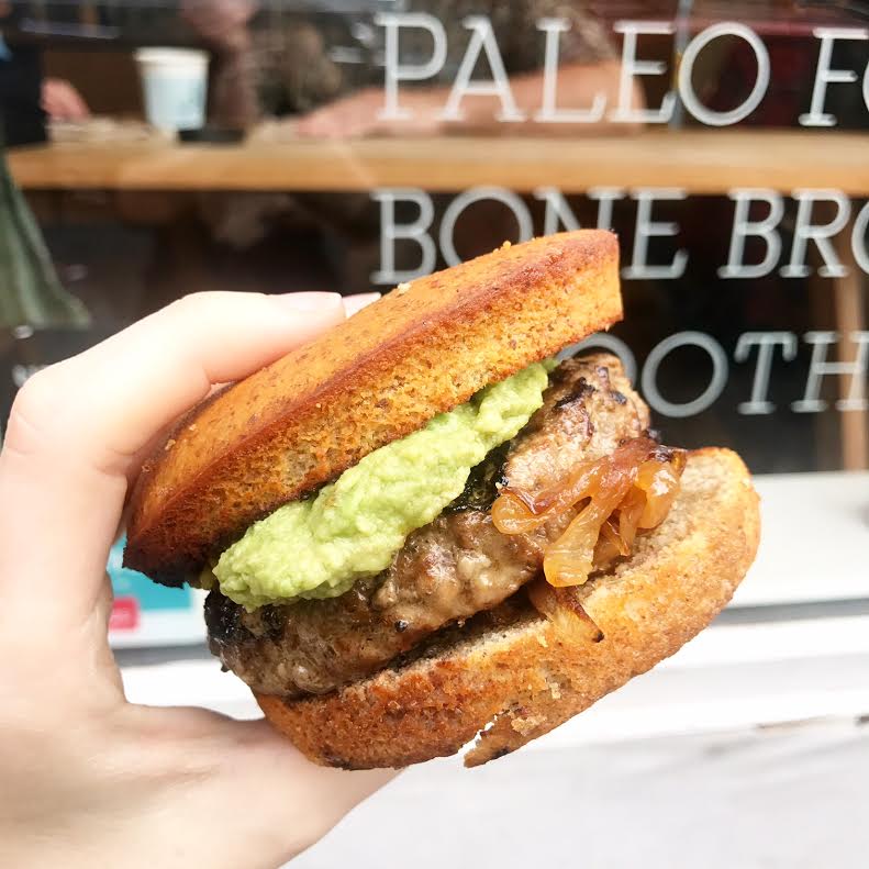 A hand holding a hamburger with avocado and a bun in front of a window.