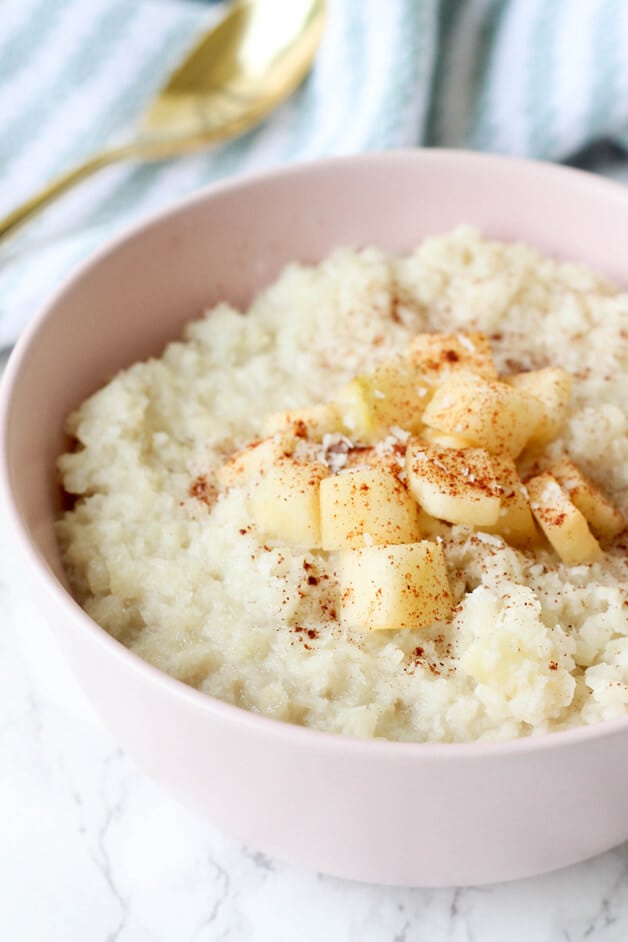 A pink bowl filled with cauliflower oats topped with cinnamon and apples.