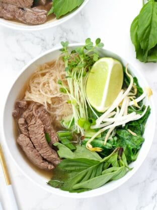 A white bowl filled with beef pho with rice noodles, herbs, lime, and meat.