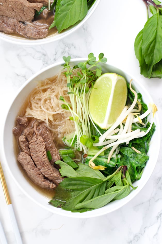 A white bowl filled with beef pho with rice noodles, herbs, lime, and meat.