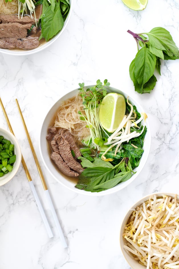 A bowl filled with pho including beef, rice noodles, herbs, bean sprouts, limes, and chop sticks next to it.