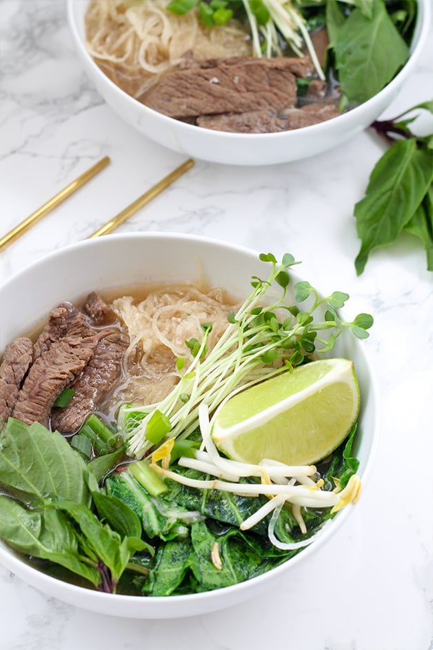 A bowl filled with pho including beef, rice noodles, herbs, bean sprouts, limes, and chop sticks next to it.