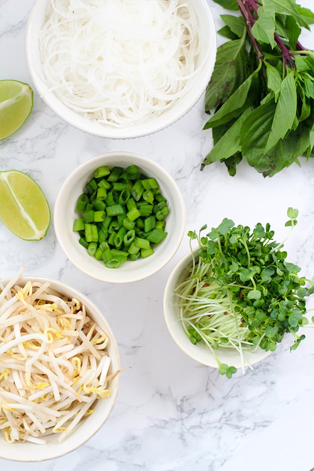 Bowls on a counter with sprouts, green onions, bean sprouts, rice noodles, and lime wedges ready to make pho.
