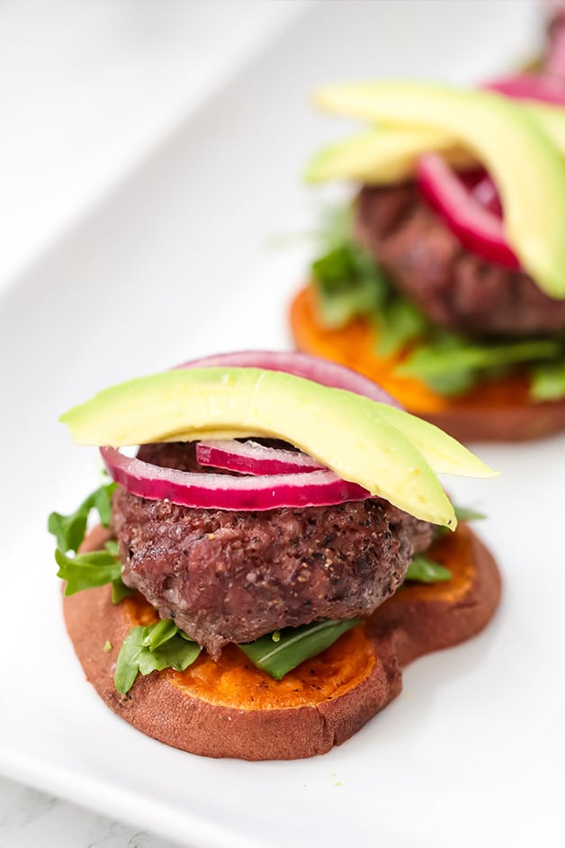 A slider on a sweet potato bun topped with red onion and avocado with another in the background.