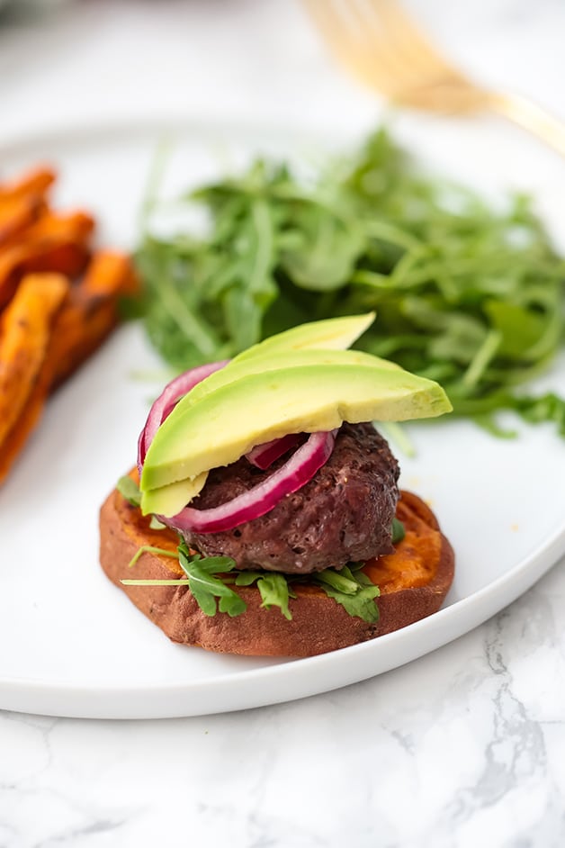 A white plate with a a beef slider on a roasted sweet potato bun toped with onion and avocado next to roasted sweet potato fries and arugula.