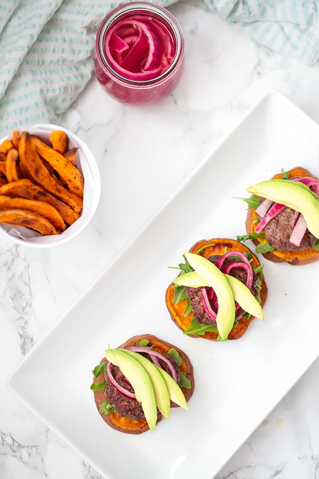 A white plate with three sliders on sweet potato buns with a bowl of sweet potato fries next to it.
