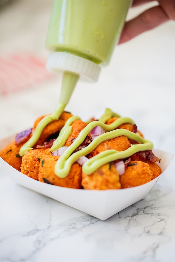 A paper bowl filled with sweet potato tots being topped with a green sauce out of a bottle.