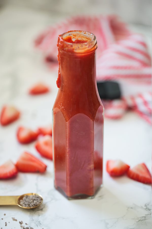 A glass bottle of strawberry balsamic BBQ suace on a counter with cut up strawberries around it.