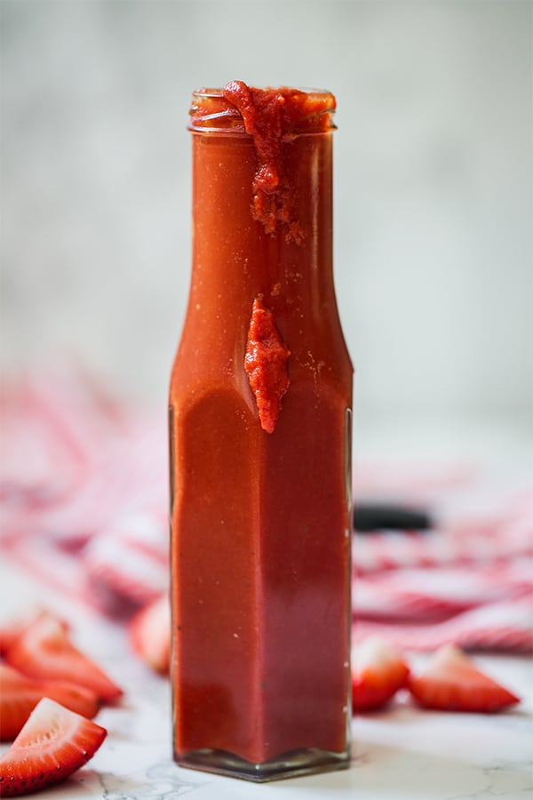 A glass bottle of strawberry balsamic BBQ suace on a counter with cut up strawberries around it.