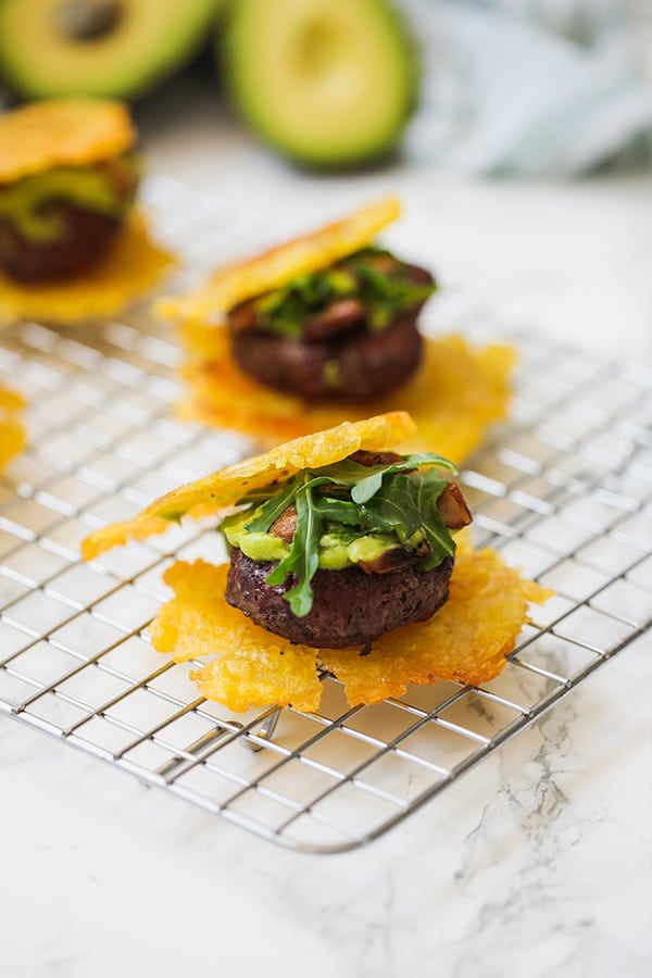 Two sliders on plantain tostone buns with greens and avocado topping sitting on a metal cooling rack.