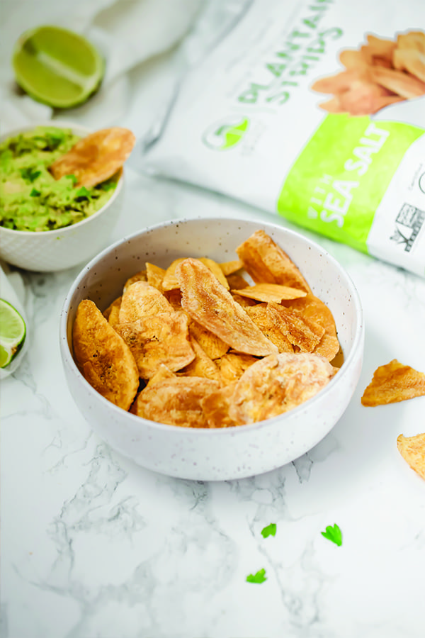A white bowl filled with plantain chips next to a smaller bowl with guacamole and one plantain chip dipping into it.