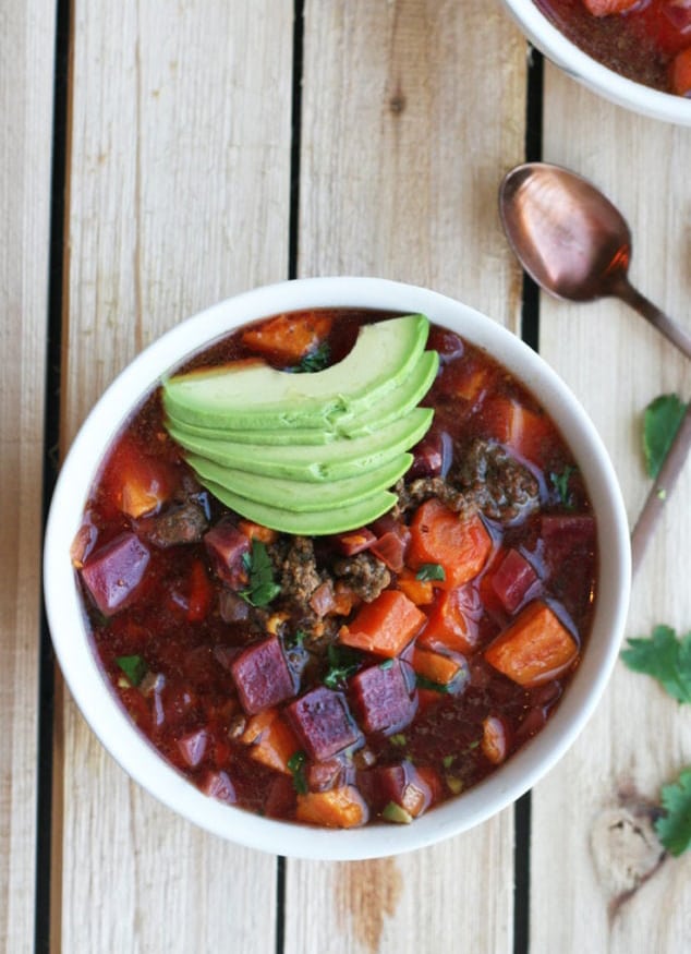 A white bowl of aip chili topped with sliced avocado on a wooden table.