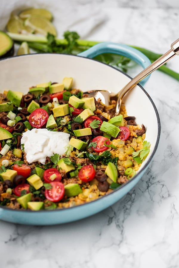 A serving dish filled with one pan taco skillet topped with avocado and coconut cream with a serving spoon in it.