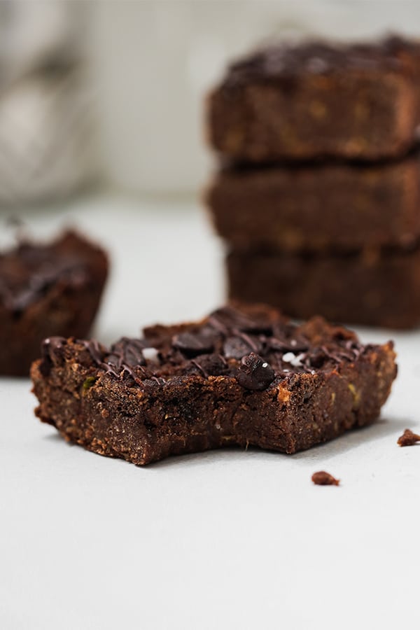 A zucchini brownie with a bite taken out of it in front of a stack of zucchini brownies.
