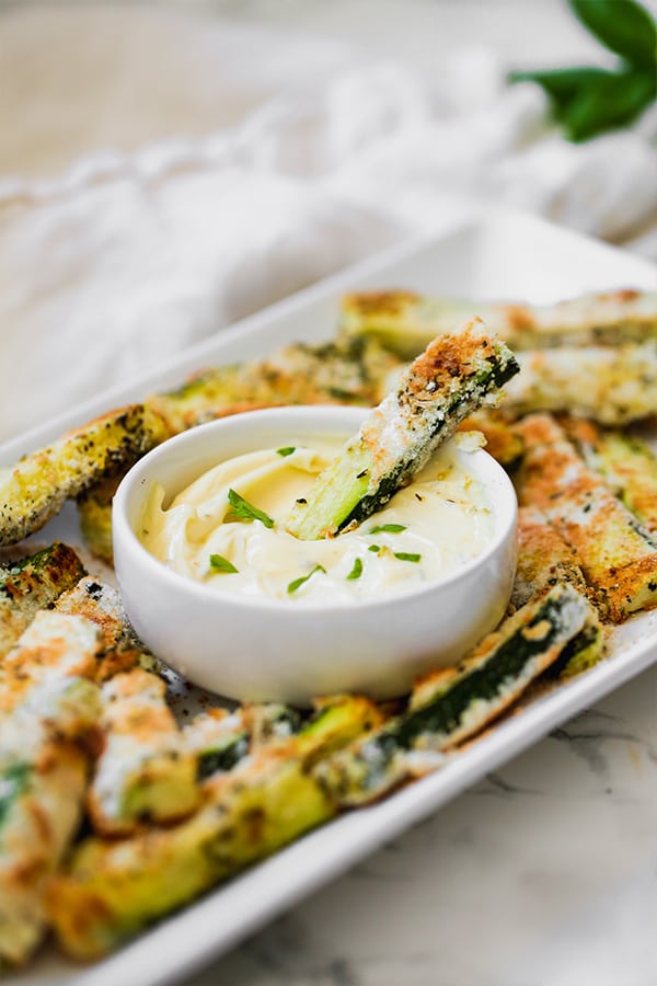 A white serving dish of zucchini fries around a white bowl of dipping sauce with one zucchini fry dipping into the sauce.