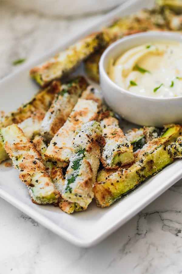 A white serving dish of zucchini fries around a white bowl of dipping sauce.