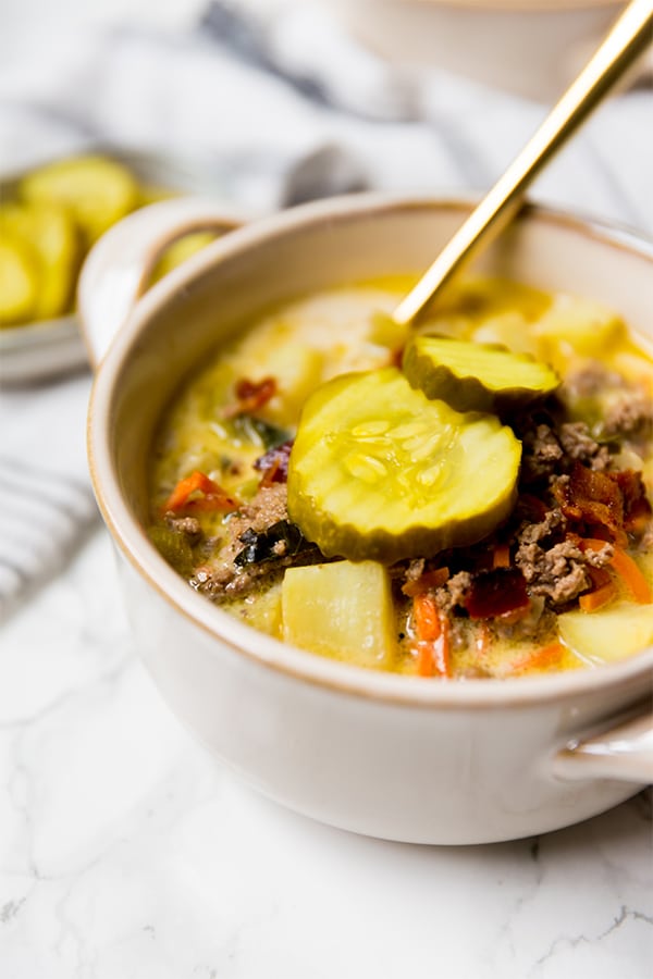 A bowl of bacon cheeseburger soup topped with pickles and with a spoon in it.