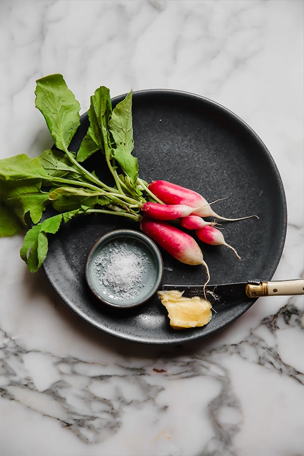 A black plate with radishes on it next to a small dish of salt and a knife.