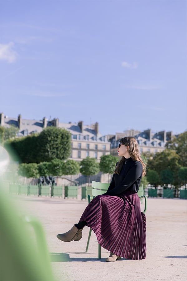 Food blogger Michelle Hoover in paris in a purple skirt sitting on a chair in a park.