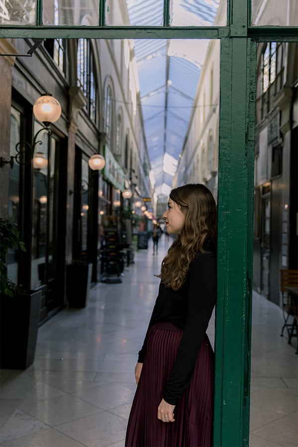 Food blogger Michelle Hoover in paris in a purple skirt and black chair standing in a shopping center leaning on a green pilar.