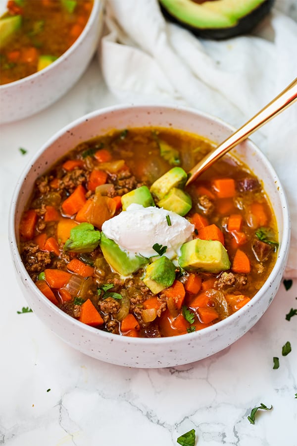 A white bowl filled with pumpkin chili topped with coconut cream and sliced avocado.