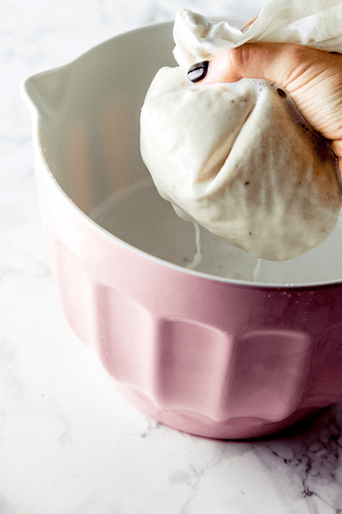 A hand holding a cheesecloth squeezing out liquid over a pink mixing bowl.