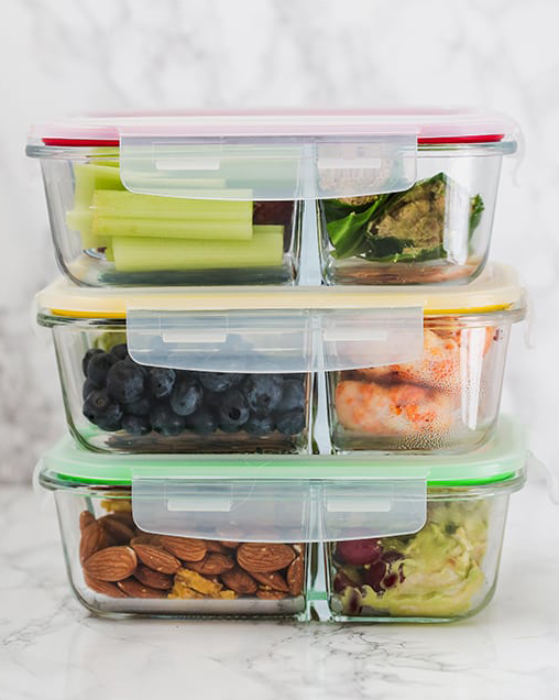 Three glass tupperwares filled with cut up fruit and vegetables stacked on top of each other.