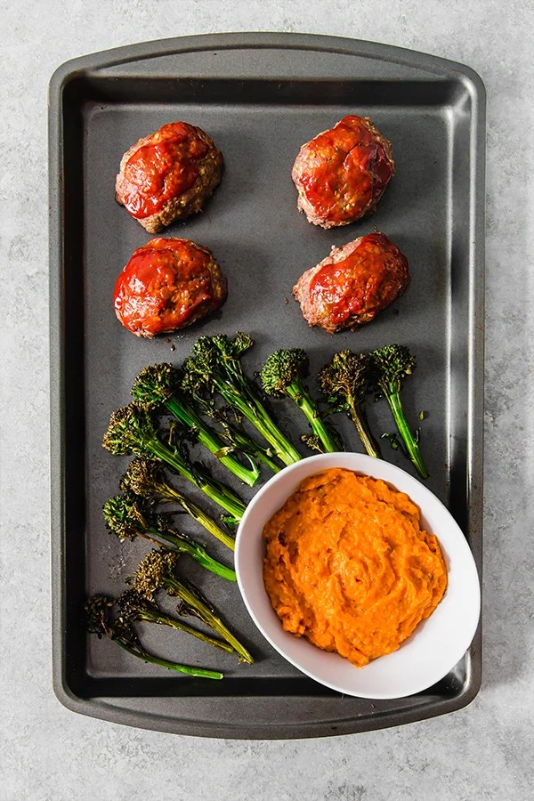 A sheet pan with roasted broccolini, mini meatloafs, and a white bowl of mashed sweet potatoes.