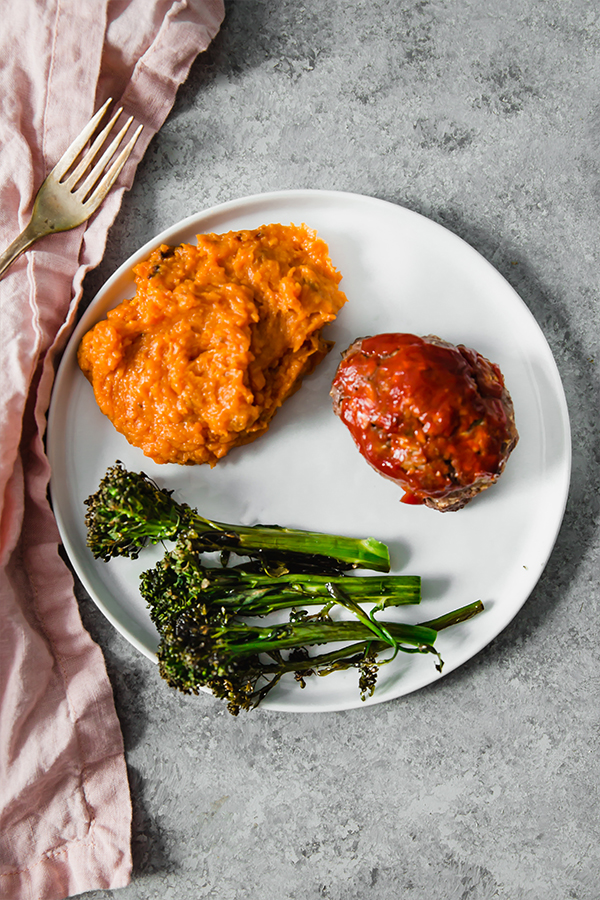 A white plate with mashed sweet potato, roasted broccolini, and slice of meatloaf on it.