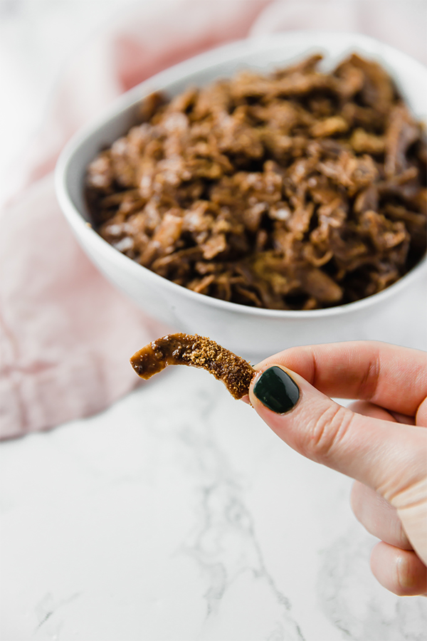 A white bowl filled with paleo coconut puppy chow with a hand holding one piece of puppy chow out in front.