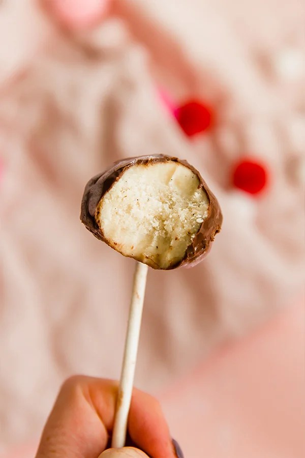 A cake pop with a bite taken out of it being held up against a fuzzy pink background.