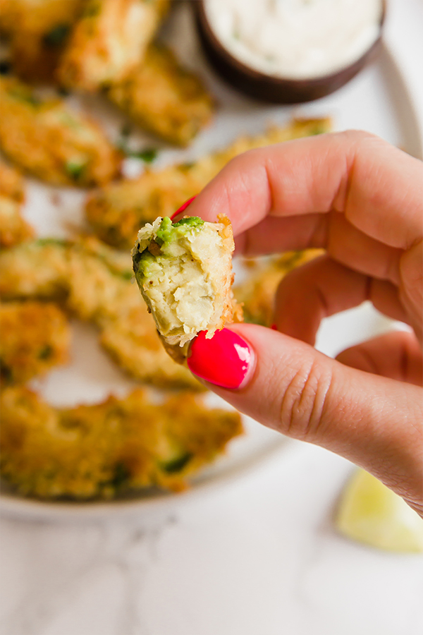 A white plate topped with avocado sections that have been coated and cooked to make avocado fries with a hand holding out one fry that has a bite taken out of it.
