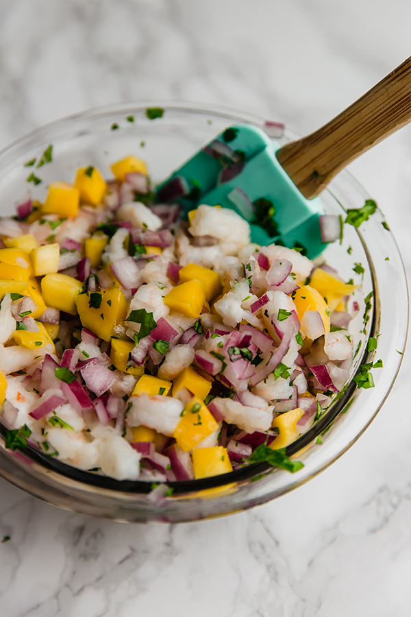 A glass bowl of the mango shrimp ceviche being mixed by a spatula.