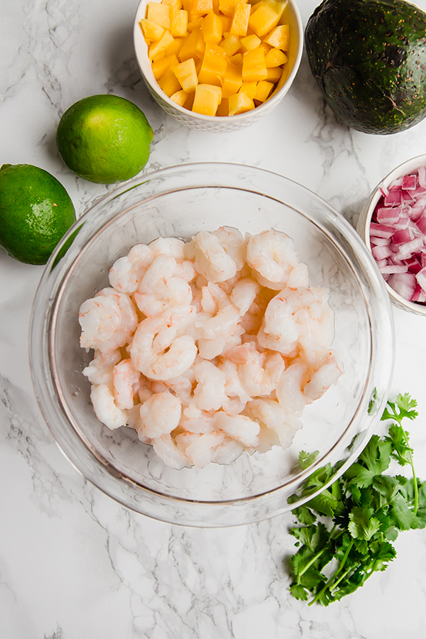 A glass bowl filled with raw shrimp surrounded by onions, cilantro, and limes.