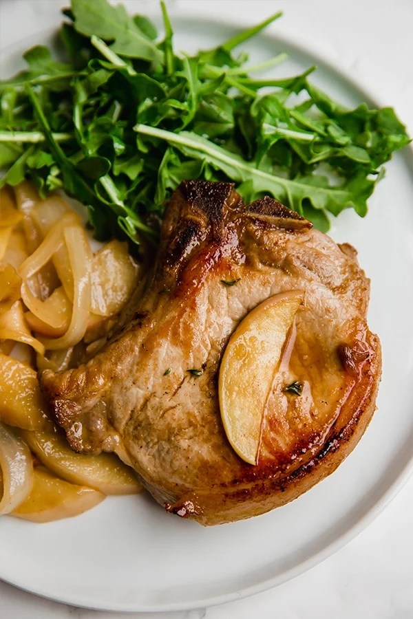 A white plate with apple pork chop on it and an arugula salad.