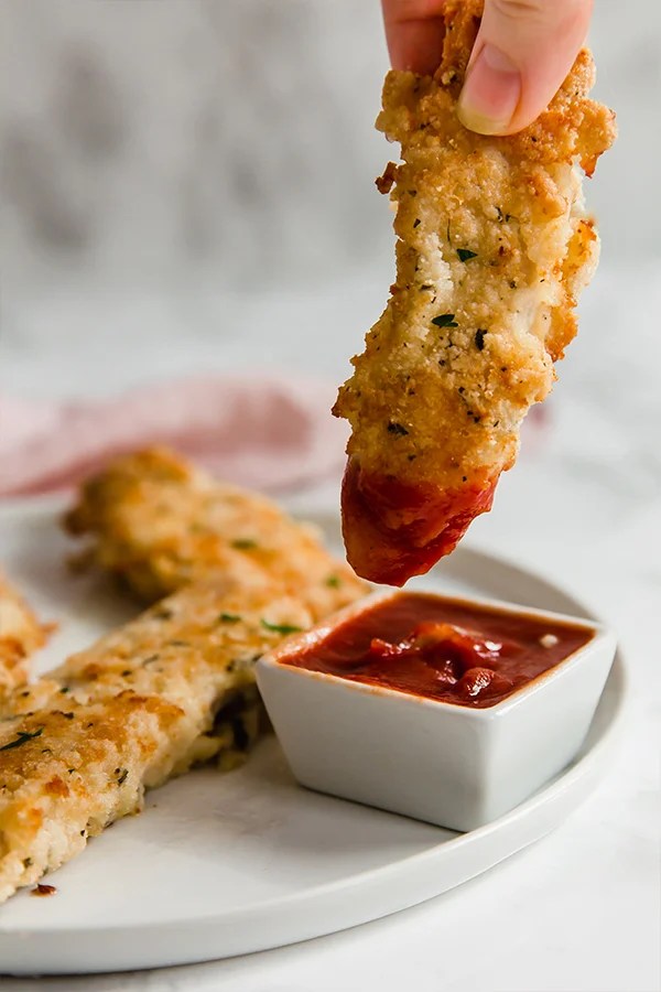 A white plate topped with homemade chicken tenders with one being dipped into a small container of dipping sauce.