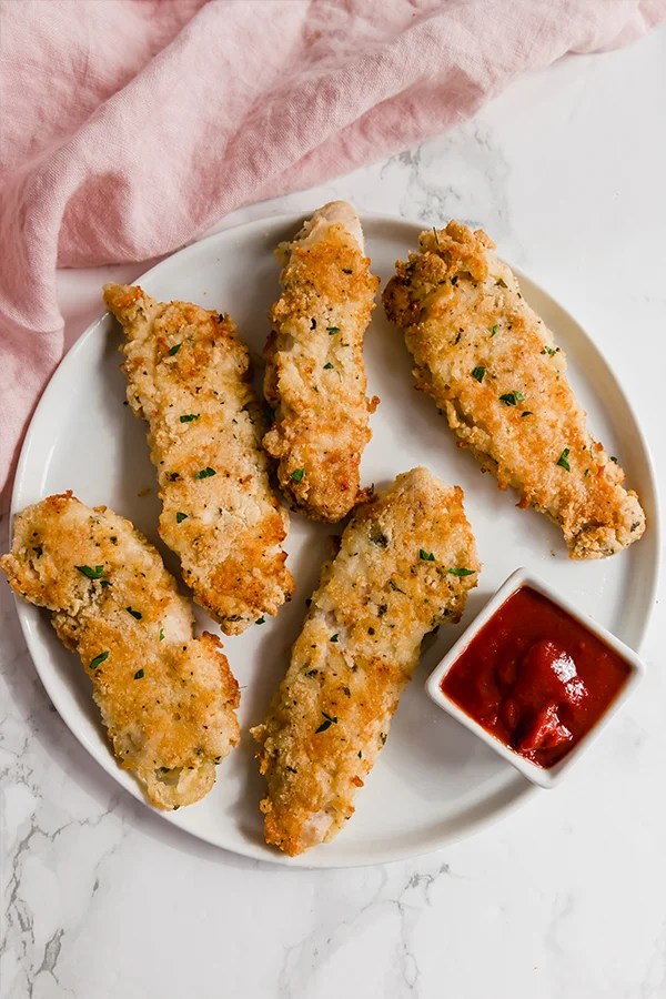 A white plate topped with homemade chicken tenders with dipping sauce in a small cup nearby.