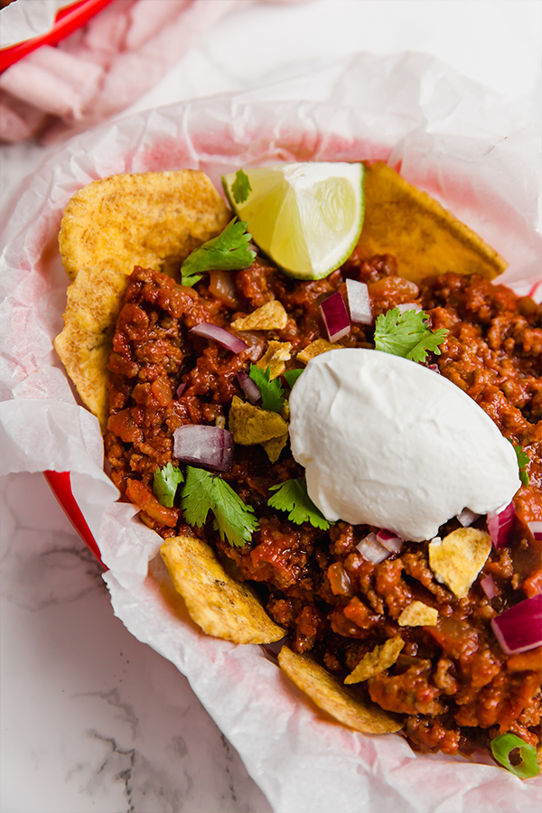 A red basket with parchment paper fille with frito pie over plantain chips with fresh lime wedges on it.