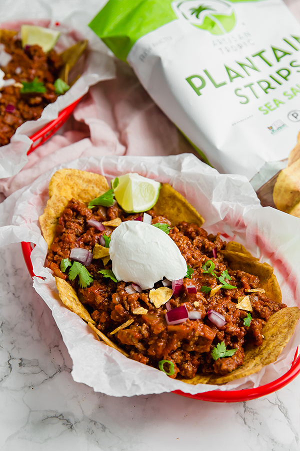 A red basket with parchment paper fille with frito pie over plantain chips with fresh lime wedges on it.