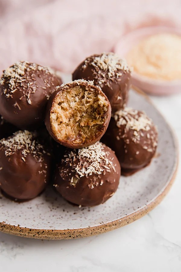 Chocolate covered samoa truffles on a small plate topped off with lightly toasted coconut, with a bite being taken out of one to show the soft coconut interior.