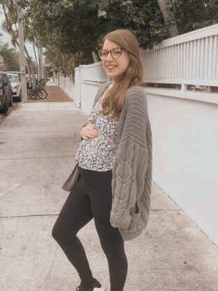 Michelle pregnant standing on a sidewalk smiling at the camera and holding her belly.