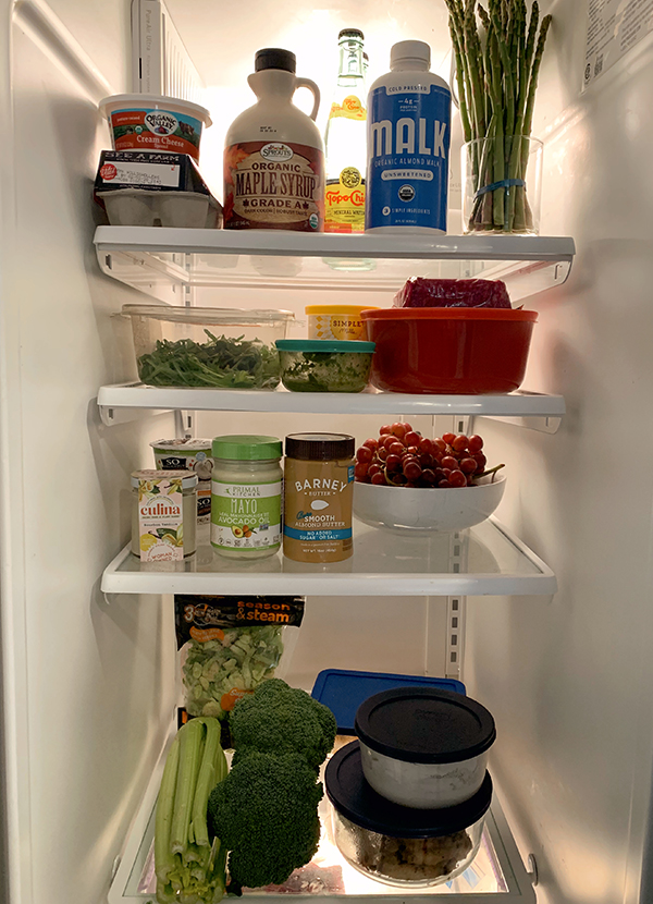 A look into the fridge with vegetables, almond butter, mayo, grapes, tupperware containers, almond milk, and maple syrup.