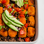 A white baking dish filled with sweet potato tater tot taco casserole topped with avocado slices.