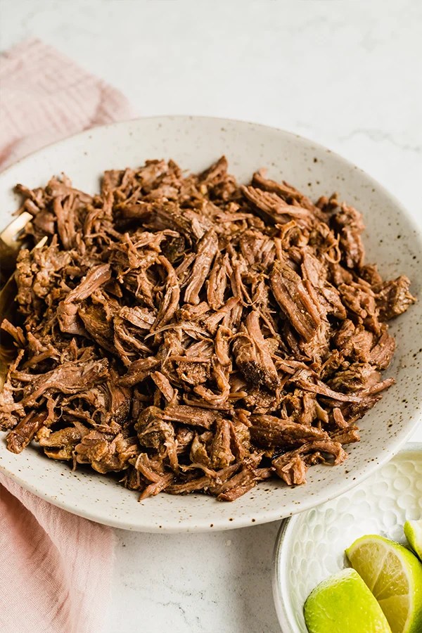 Shredded slow cooker beef in a bowl with a pink kitchen towel in the background.