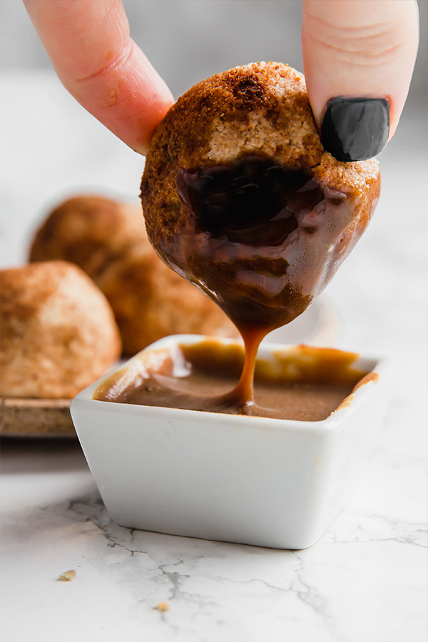 A hand dipping a dulce de leche filled donut hole into sauce.