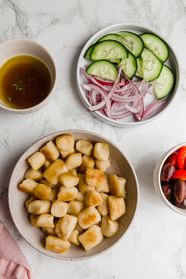 All the ingredients needed to make Greek gnocchi salad laid out in bowls on a counter.