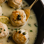 Chicken piccata meatballs in sauce in a pan with a spoon scooping out one meatball.